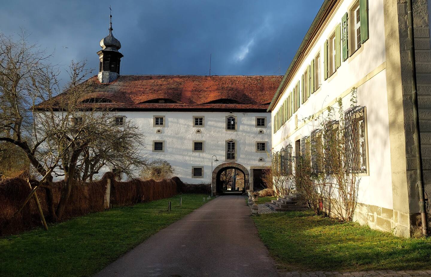 Eine Teilaufnahme des herrschaftlichen Schlosses von außen. Es ist weiß verputzt, hat viele Fenster sowie einen kleinen Turm und einen Torborgen. Davor stehen Bäume in einer Grünfläche.